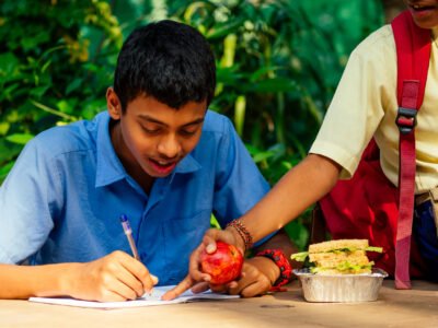 indian school boy writing on notepad doing homework , looking concentration . His friend with backpack sits on table and eating an apple in park.